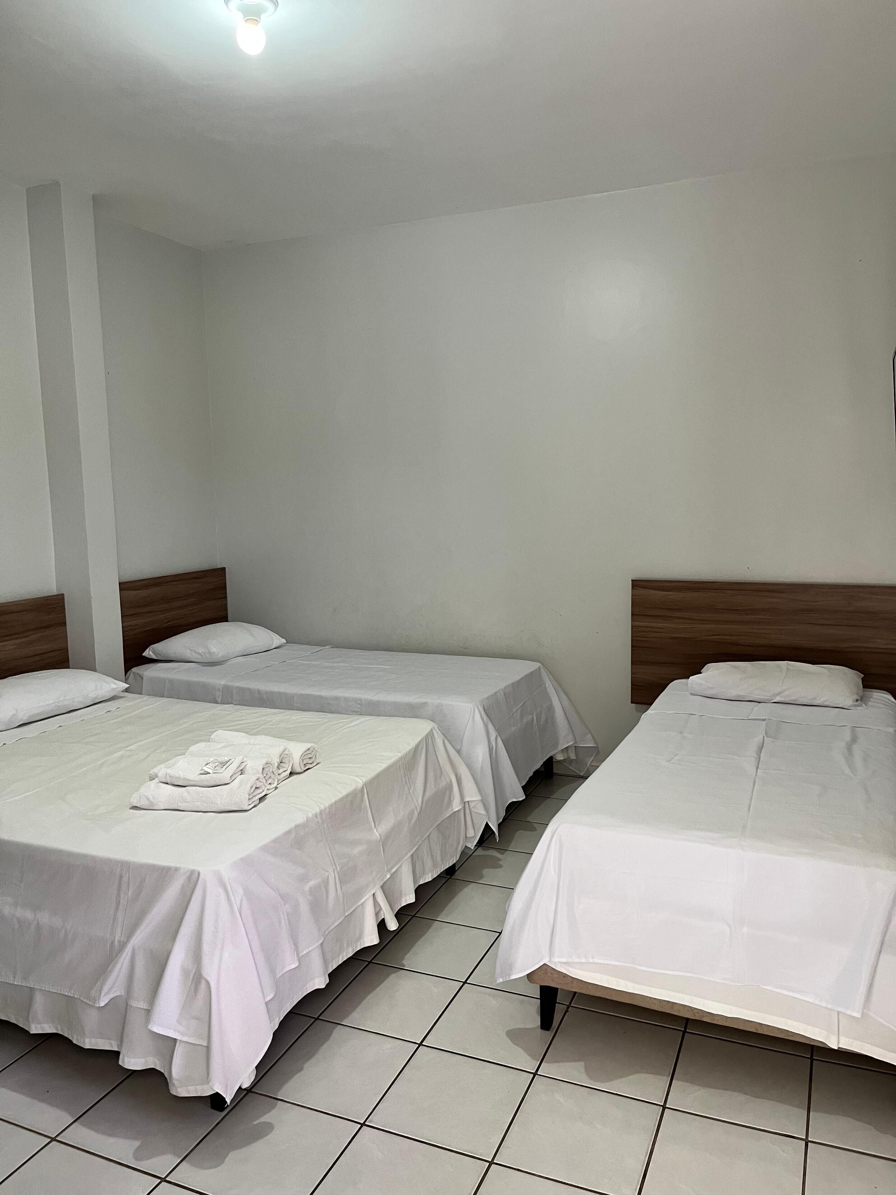 A hotel room with three single beds, each with white bedding and pillows. The room has white walls and a tiled floor.
