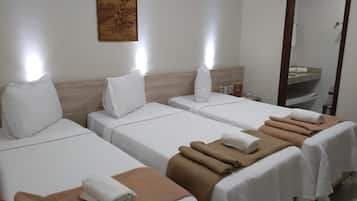 Quarto de hotel com três camas de solteiro, cada uma com um edredom branco e uma toalha marrom. Há uma cabeceira de madeira, uma obra de arte na parede e um espelho sobre a pia.