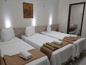 Habitación de hotel con tres camas individuales, cada una con una colcha blanca y una toalla marrón. Se observa un cabecero de madera, una obra de arte colgada en la pared y un espejo sobre el lavamanos.