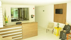 A reception area with a wooden reception desk, a glass partition, a television mounted on the wall, and a small yellow table with a plant. - Hotel Padre Cícero (Juazeiro do Norte)