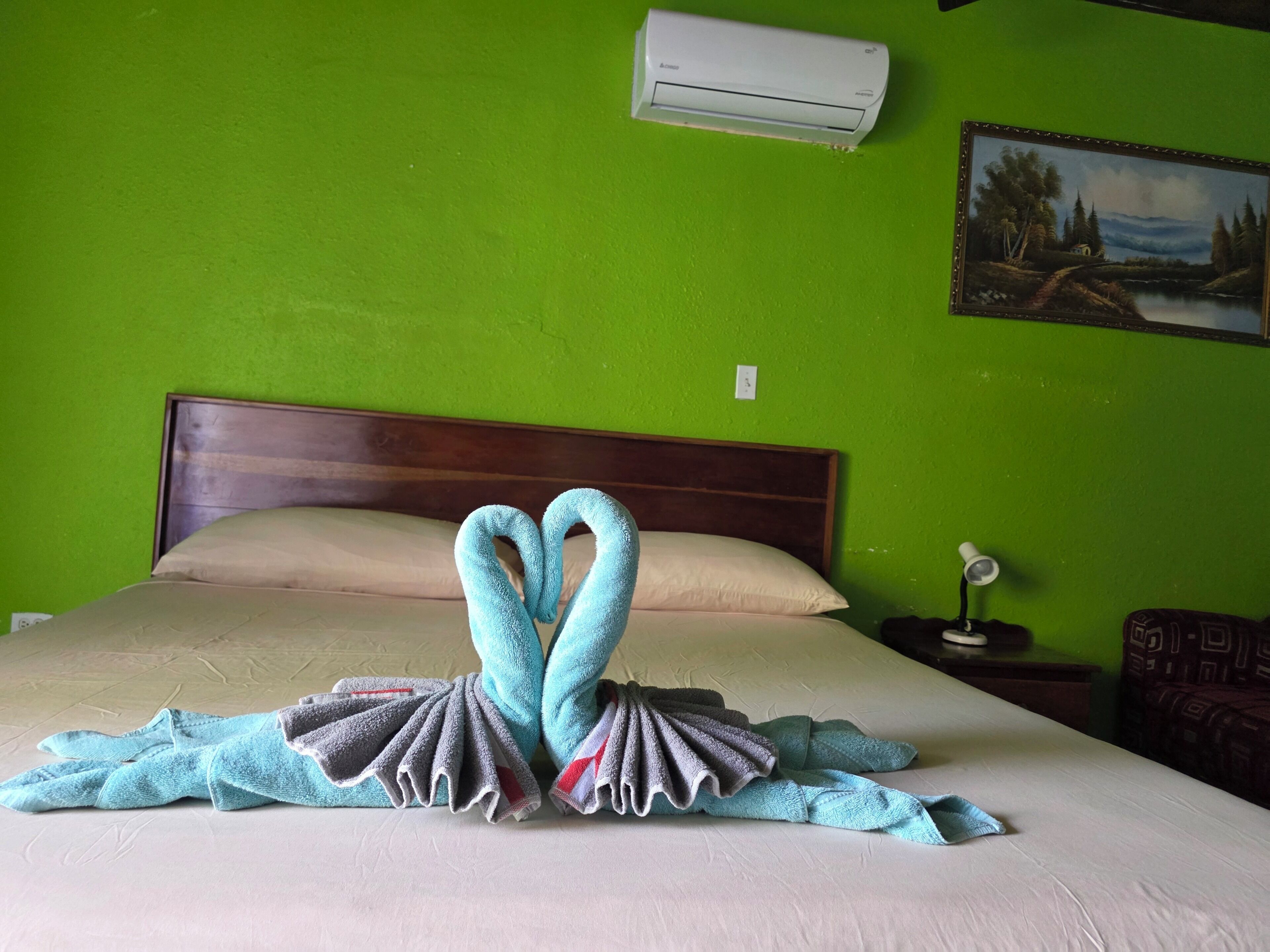 Una habitación de hotel con cama, pared verde, aire acondicionado, un cuadro y una mesita de noche con lámpara.