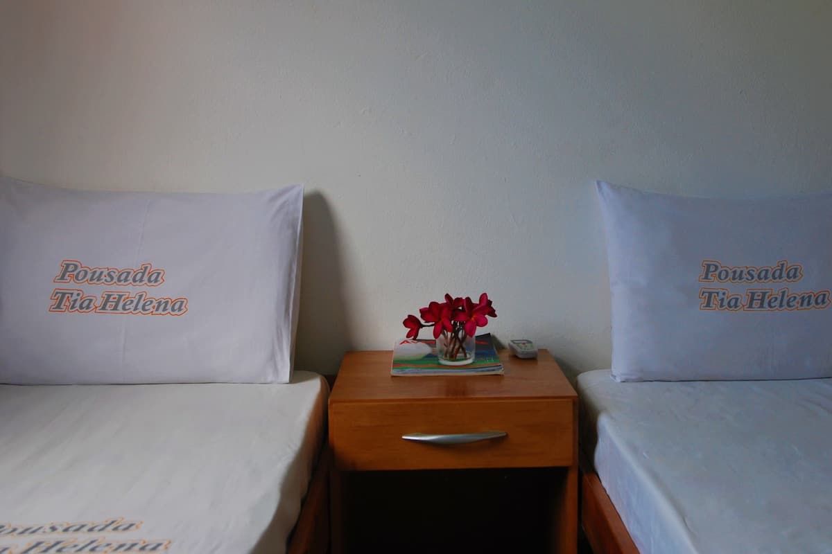 A hotel room with two beds, each with a pillow labeled "Pousada Tia Helena." A small table with a vase of red flowers is between the beds.
