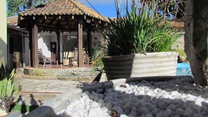 A covered outdoor seating area with a tiled roof, surrounded by potted plants and a stone pathway leading to a pool. - Pousada Villa Guimarães (Chapada dos Guimarães)