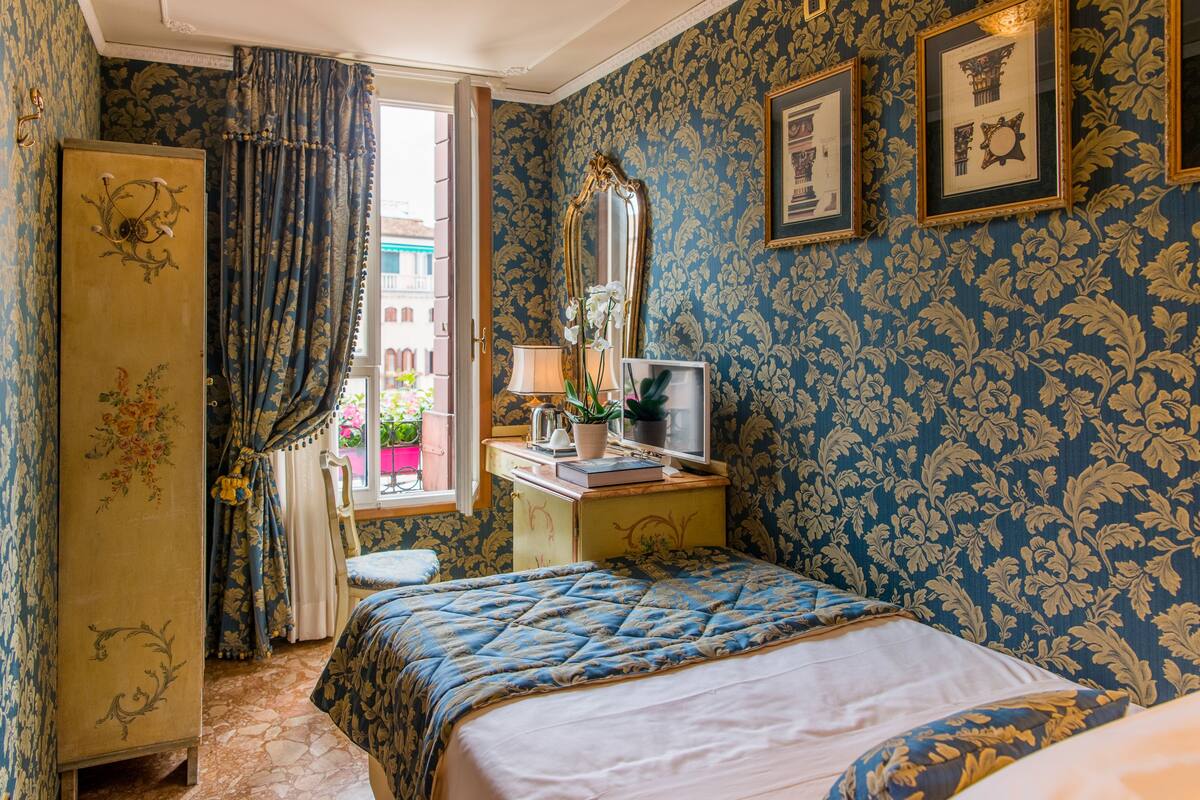 A bedroom with a bed, a desk with a computer, a mirror, and a window with curtains.