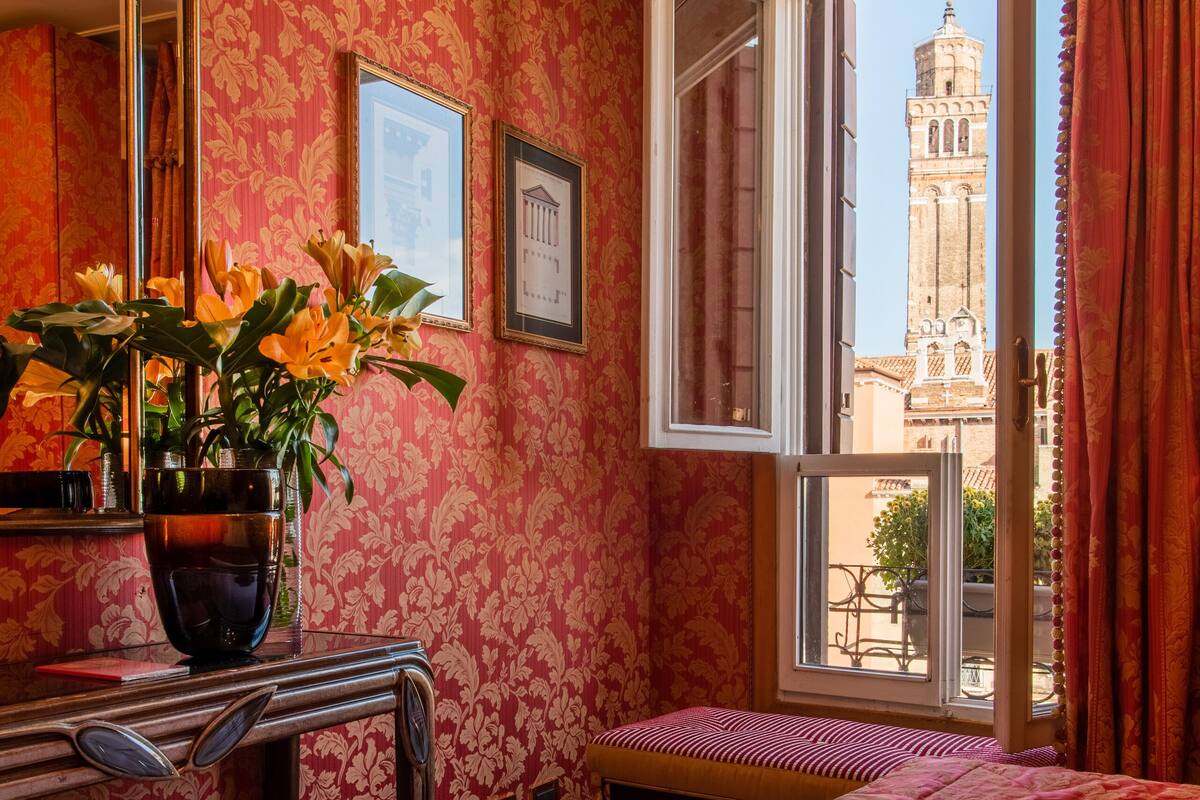 A room with a bed, a desk with a vase of flowers, a window with a view of a tower, and patterned wallpaper.