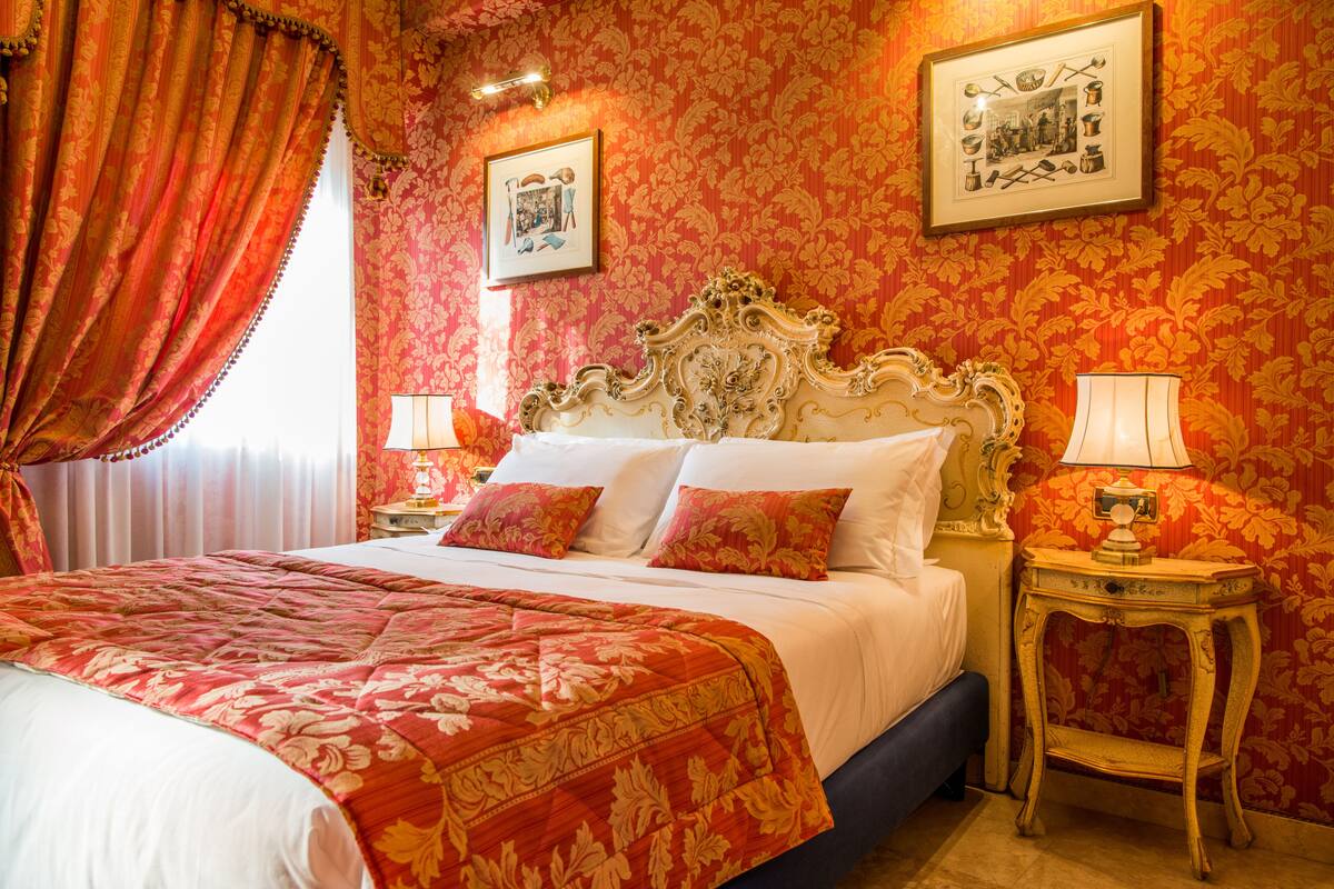 A hotel room with a large bed, ornate headboard, floral wallpaper, and two bedside tables with lamps.