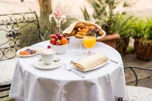 Una mesa redonda con mantel blanco, un plato de croissants, un bol de fruta, una taza de café, un vaso de zumo de naranja y una servilleta doblada.