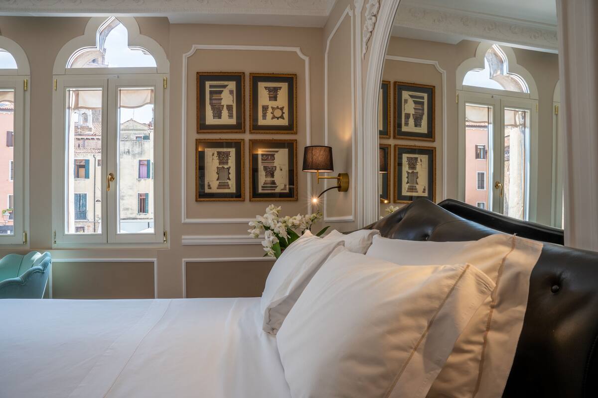 A hotel room with a bed, a lamp, framed pictures on the wall, and windows with curtains.