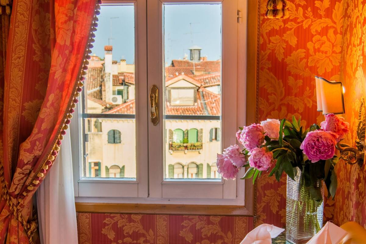 A hotel room with a bed, floral curtains, a vase with flowers, and a view of buildings through a window.