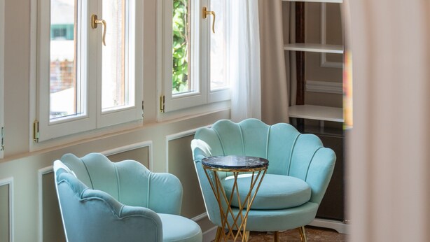 A room with two light blue armchairs, a small round table, and a white bed. There are two large windows with curtains and a white ceiling with decorative molding. - Hotel Santo Stefano (Venice)