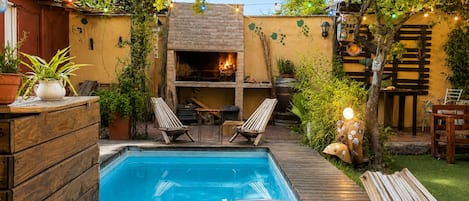 A backyard with a swimming pool, a wooden deck, and a brick fireplace.