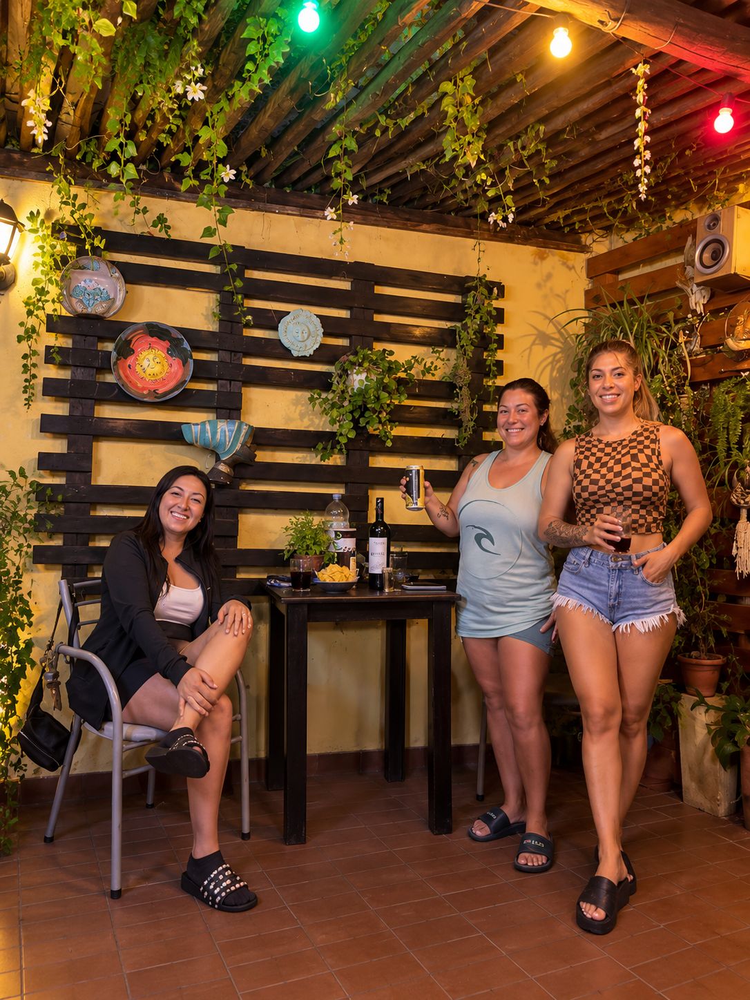 Un groupe de personnes se trouve dans un cadre extérieur rustique, agrémenté de poutres en bois, de plantes en pot et d’une table où sont disposées des boissons.