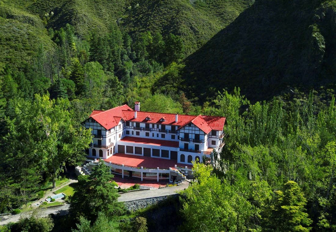 A large, multi-story building with a red roof, surrounded by dense forest and hills.