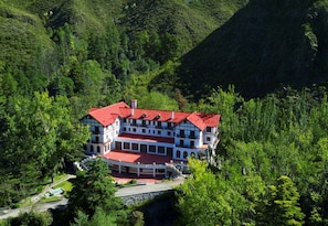 Un grand immeuble à plusieurs étages, coiffé d’un toit rouge, entouré d’une forêt dense et de collines.