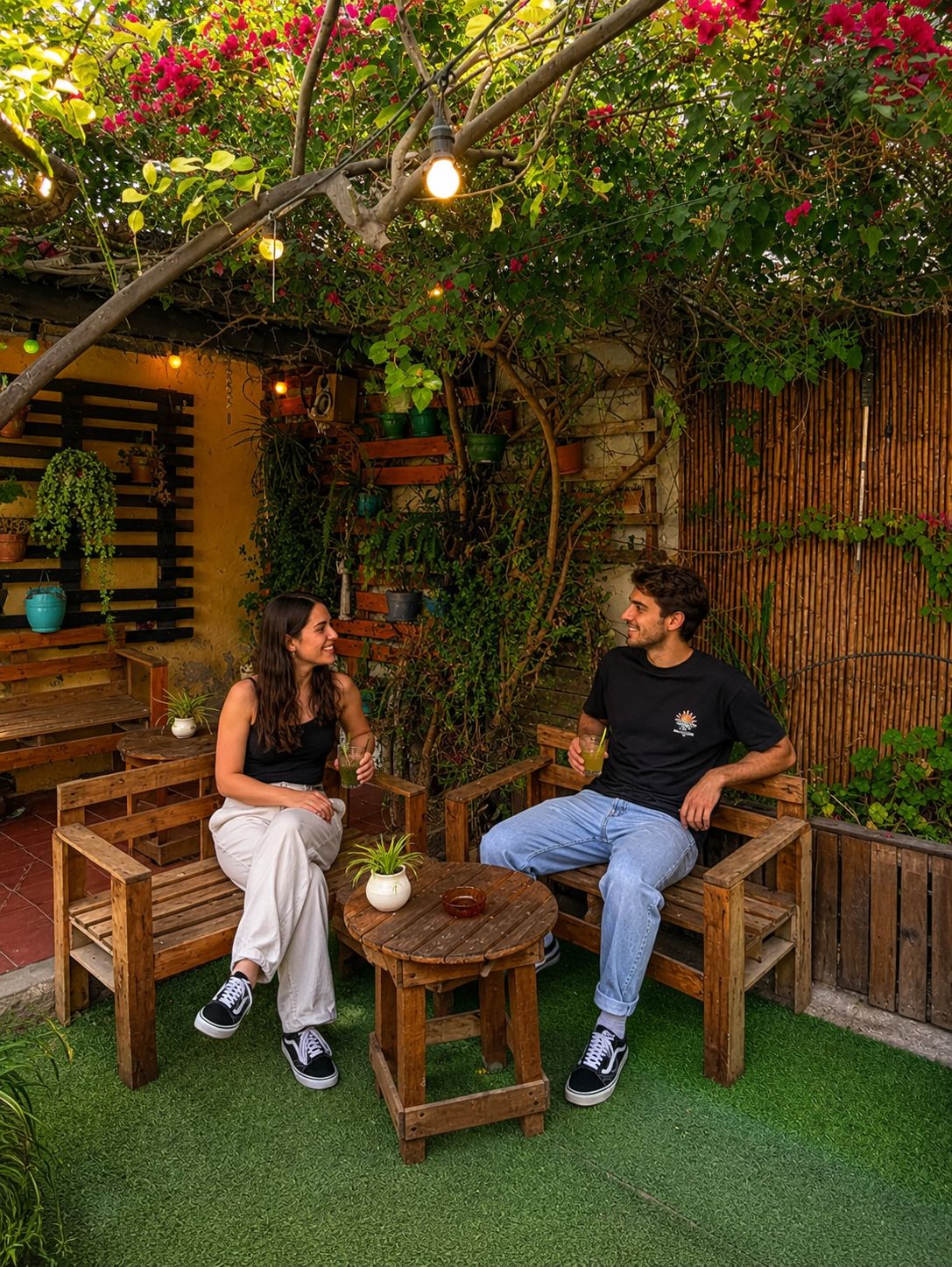 Una pareja sentada en bancos de madera en un jardín con césped artificial, rodeada de plantas en macetas y una valla de bambú.