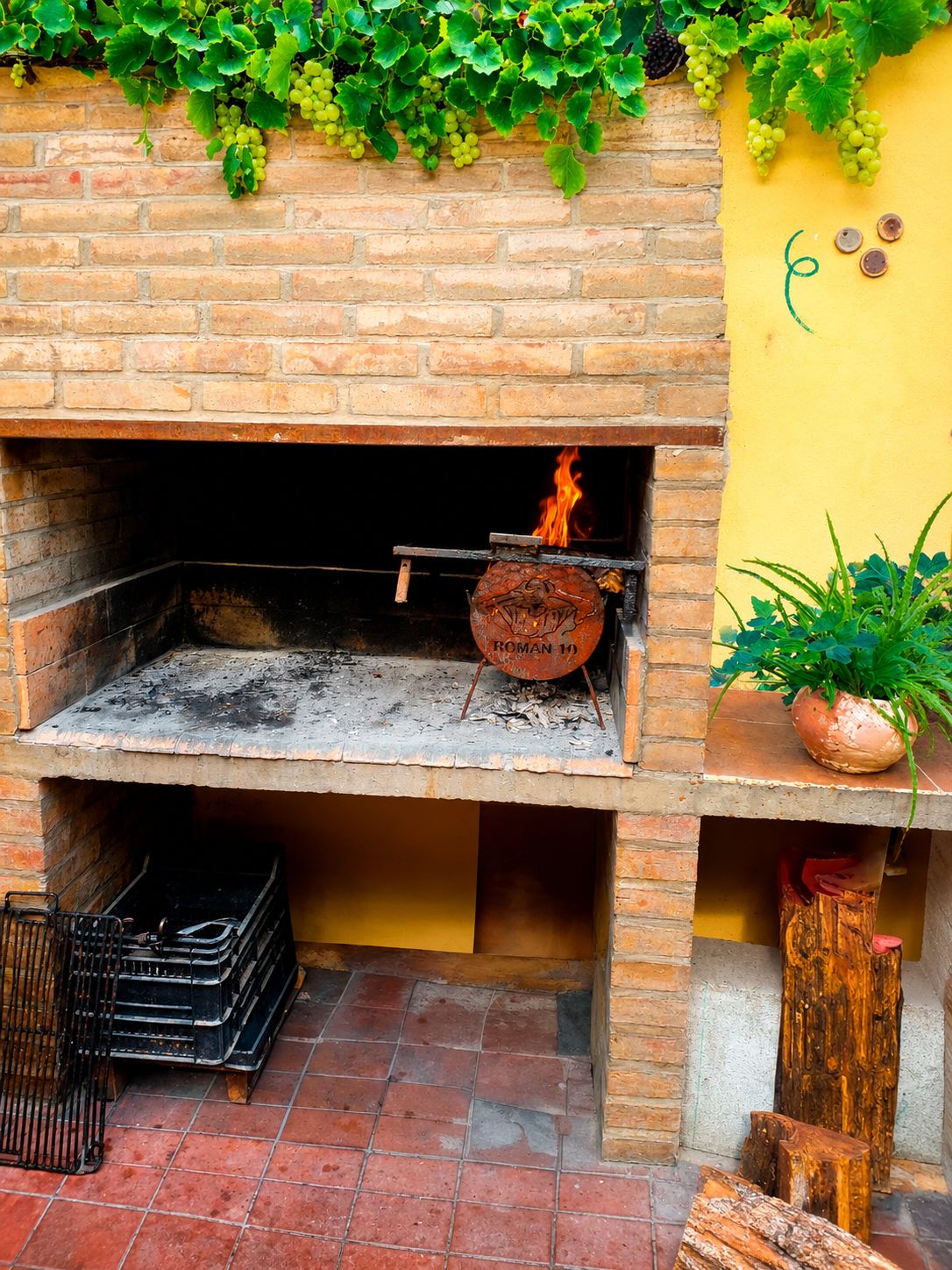 Un horno de ladrillo al aire libre con fuego encendido, rodeado por una pared amarilla y follaje.