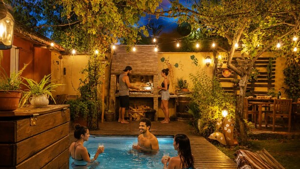 A swimming pool area with a wooden deck, a table with snacks, and string lights. - Estacion Mendoza Hostel (Mendoza)