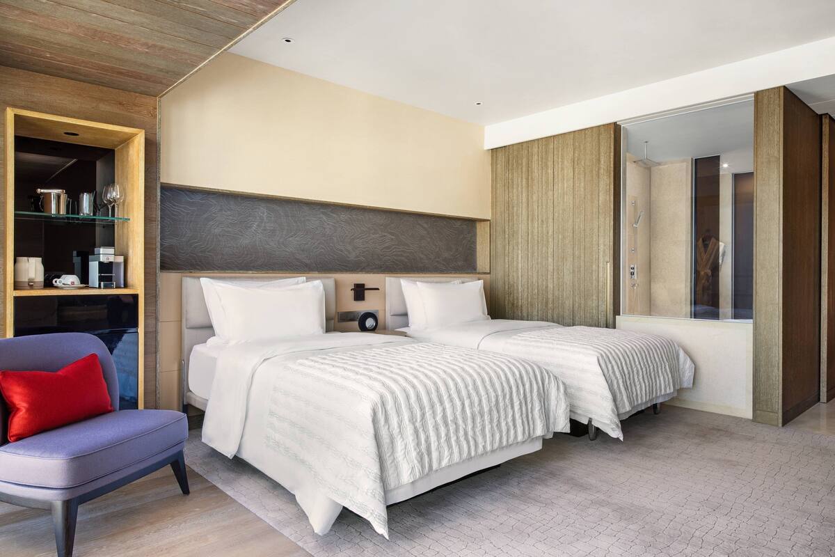 A modern bedroom with two beds, a seating area, and a wardrobe.