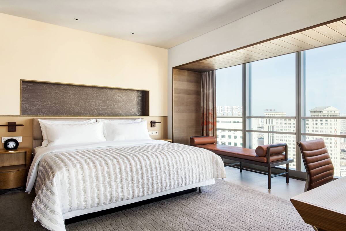 A modern hotel room with a large bed, a desk, and a view of the city through large windows.