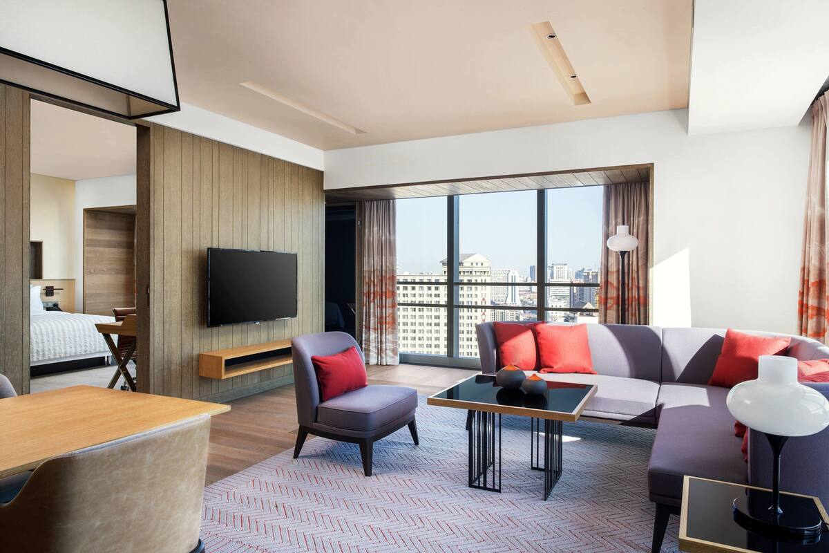 A modern hotel room with a large window, a flat-screen TV, a sofa, a chair, a coffee table, and a desk.