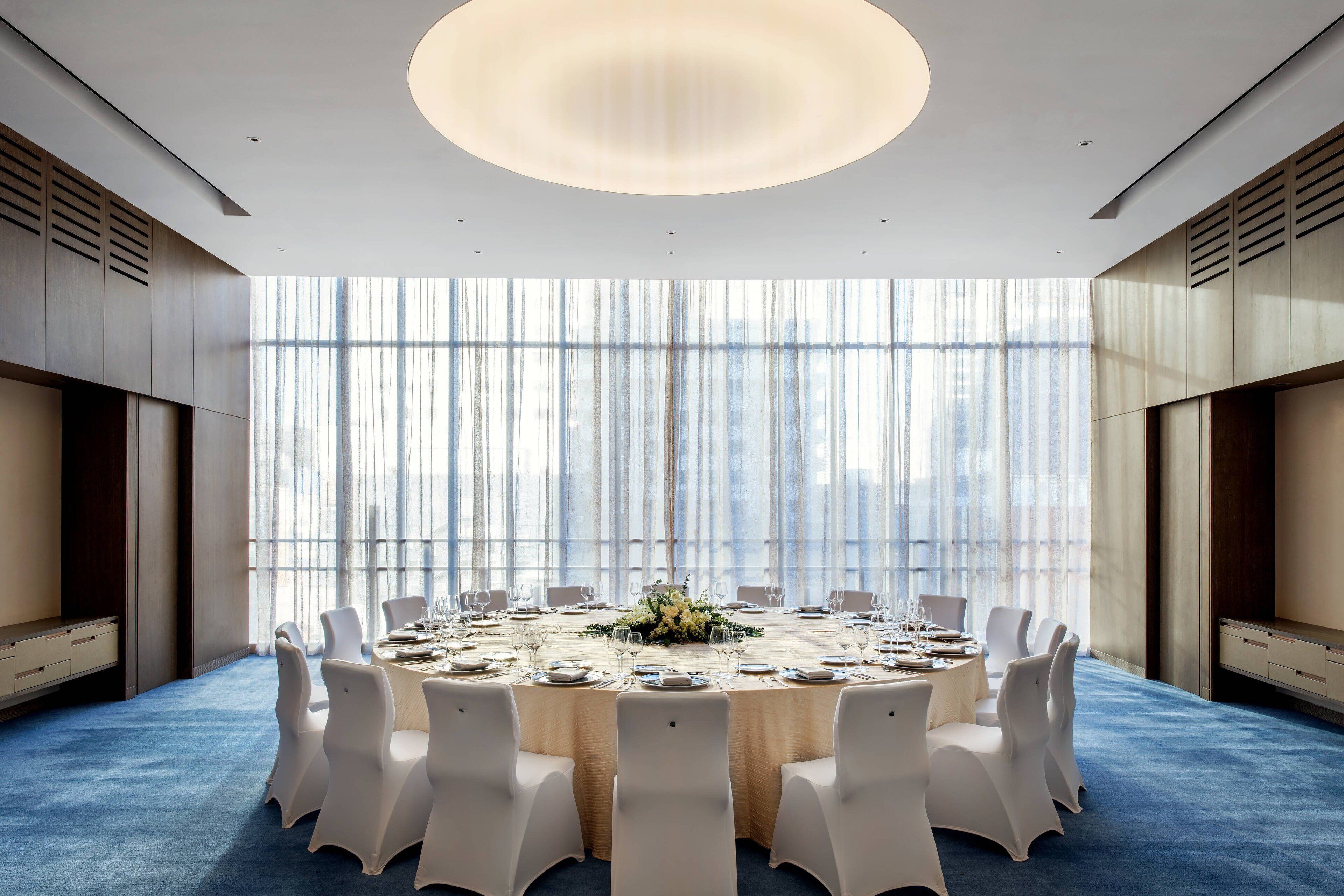 Une salle de conférence avec une table ronde, des chaises blanches et une grande fenêtre ornée de rideaux légers.