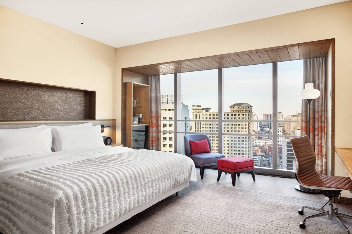 A hotel room with a large bed, a desk with a chair, a red ottoman, and a view of the city through large windows.
