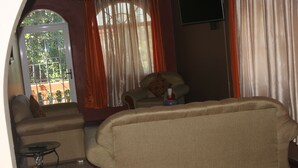 A living room with a sofa, armchair, and a small table. There is a window with curtains, a TV mounted on the wall, and a balcony visible through the window. - Cozy Apartments (Kingston)