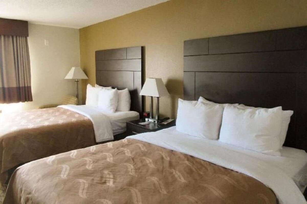 A hotel room with two beds, a nightstand with a lamp, and a headboard with a dark panel.