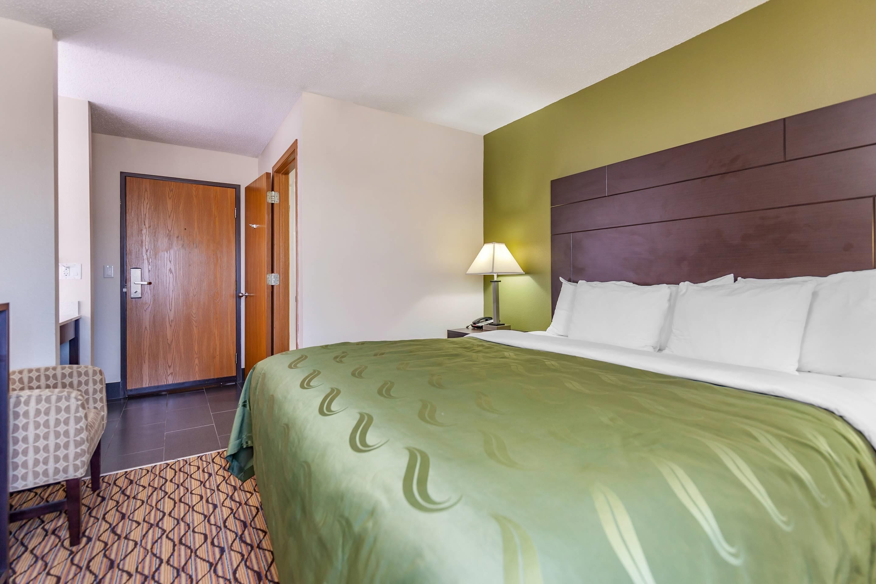 A hotel room with a large bed, a wooden door, a chair, and a lamp.