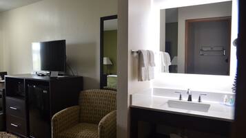 A hotel room with a television, a chair, a desk, a sink, and a mirror.