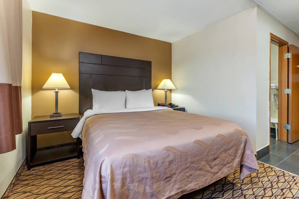Una habitación de hotel con una cama grande, dos mesitas de noche con lámparas, un cabecero de madera y una alfombra estampada.
