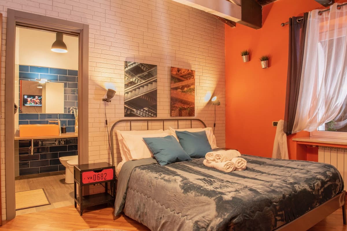 Une chambre à coucher comprenant un lit, une table de chevet, un miroir, une fenêtre avec des rideaux et un mur orné d’un tableau.