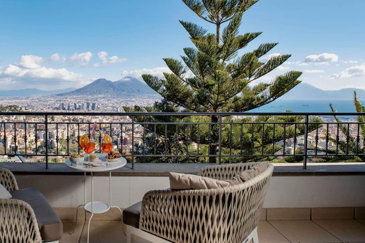 Un balcon agrémenté de meubles en osier, une petite table avec des boissons, et une vue sur la ville, comprenant des montagnes et la mer.