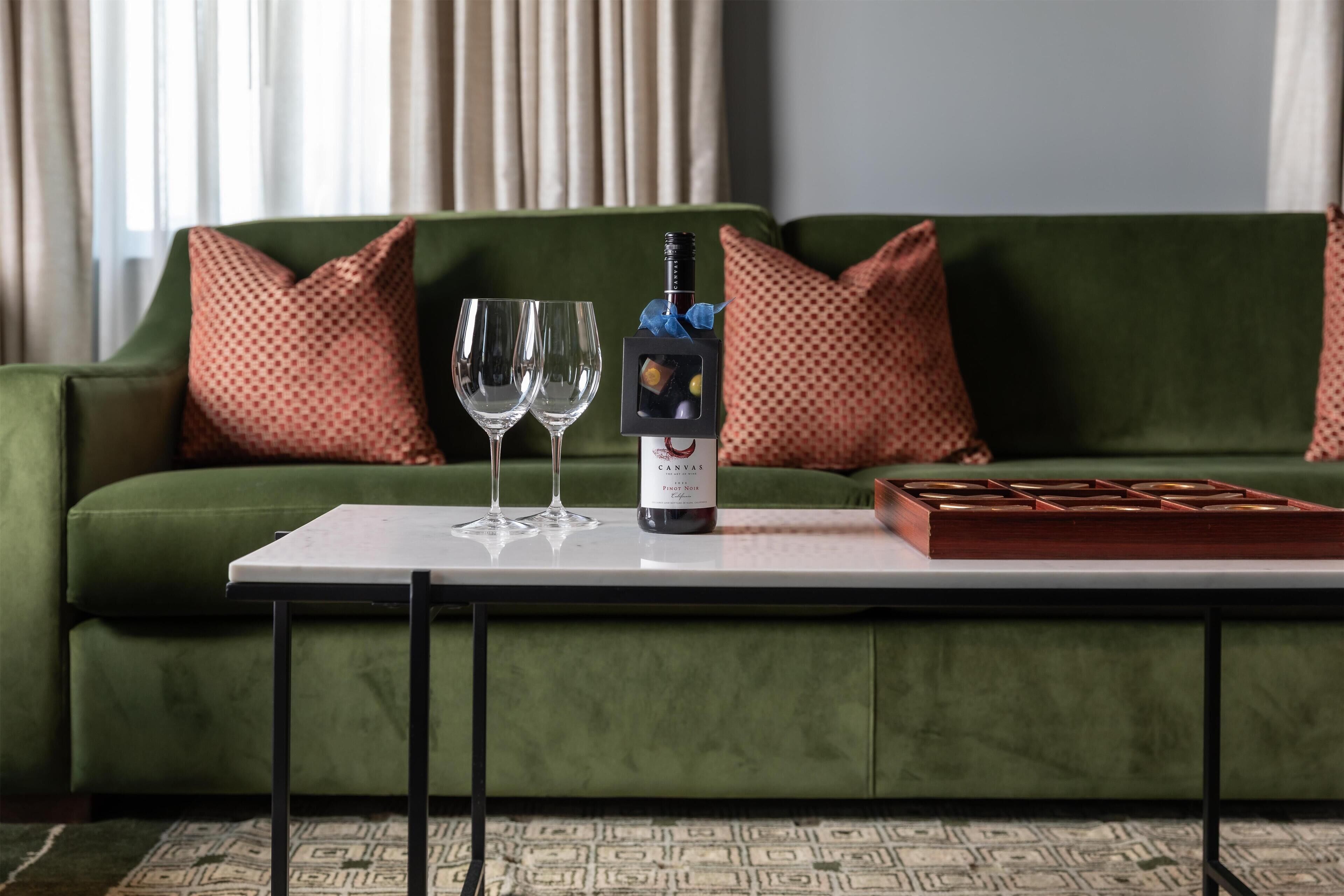 A green sofa with patterned pillows, a white table with a bottle of wine and two glasses, and a tray with a box.