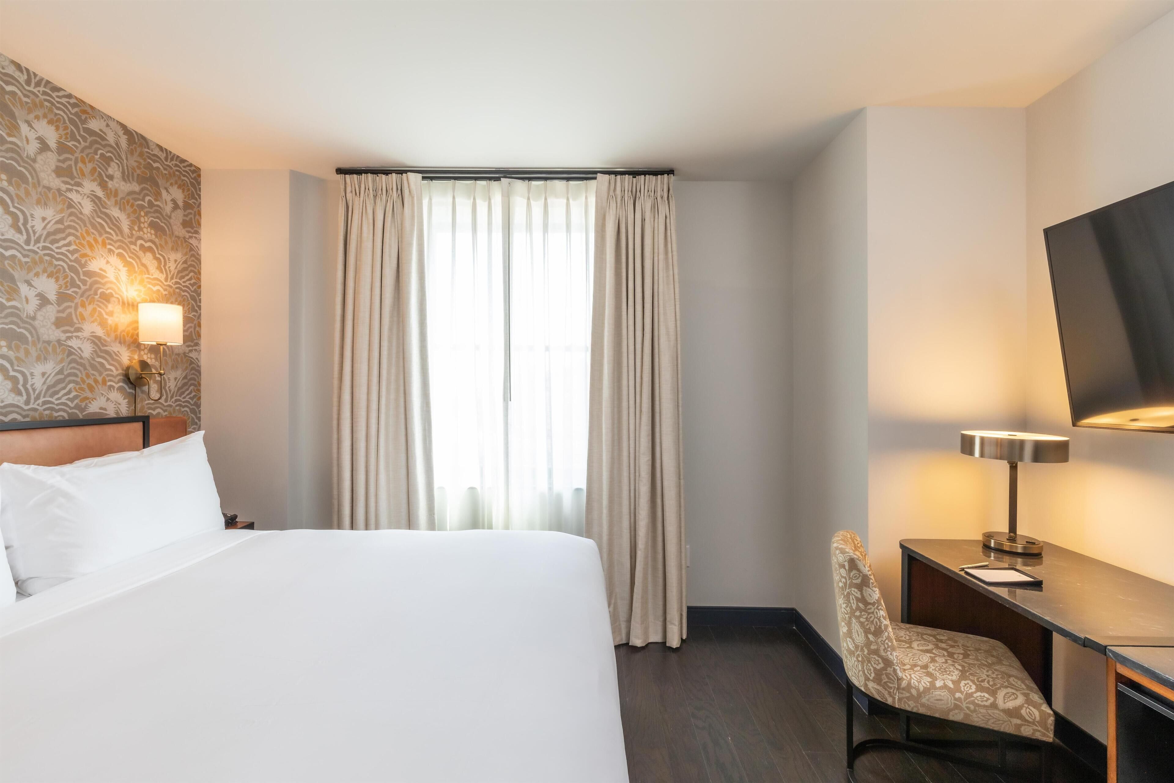 A hotel room with a large bed, a desk with a chair, a flat-screen TV, and a window with curtains.