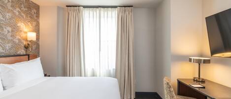 A hotel room with a large bed, a desk with a chair, a flat-screen TV, and a window with curtains.