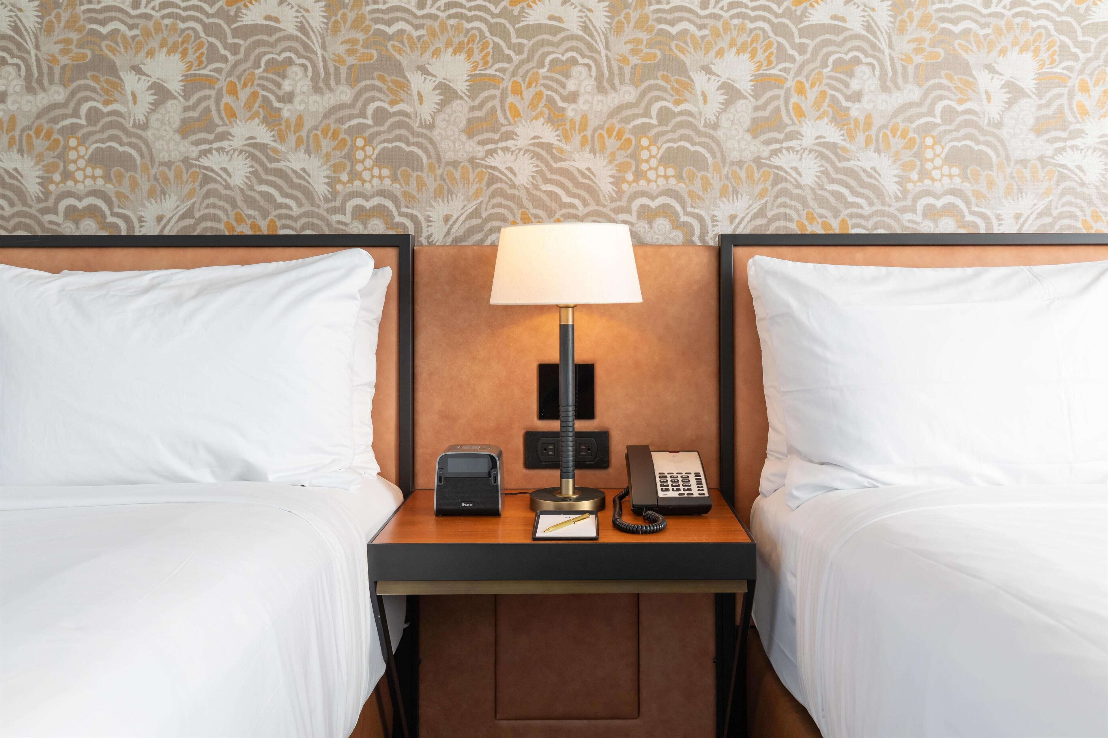 A hotel room with two beds, a nightstand with a lamp, a phone, and a small table with a sign.