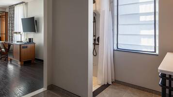 A hotel room with a wooden desk, a flat-screen TV, a shower area with a white curtain, and a large window providing natural light.