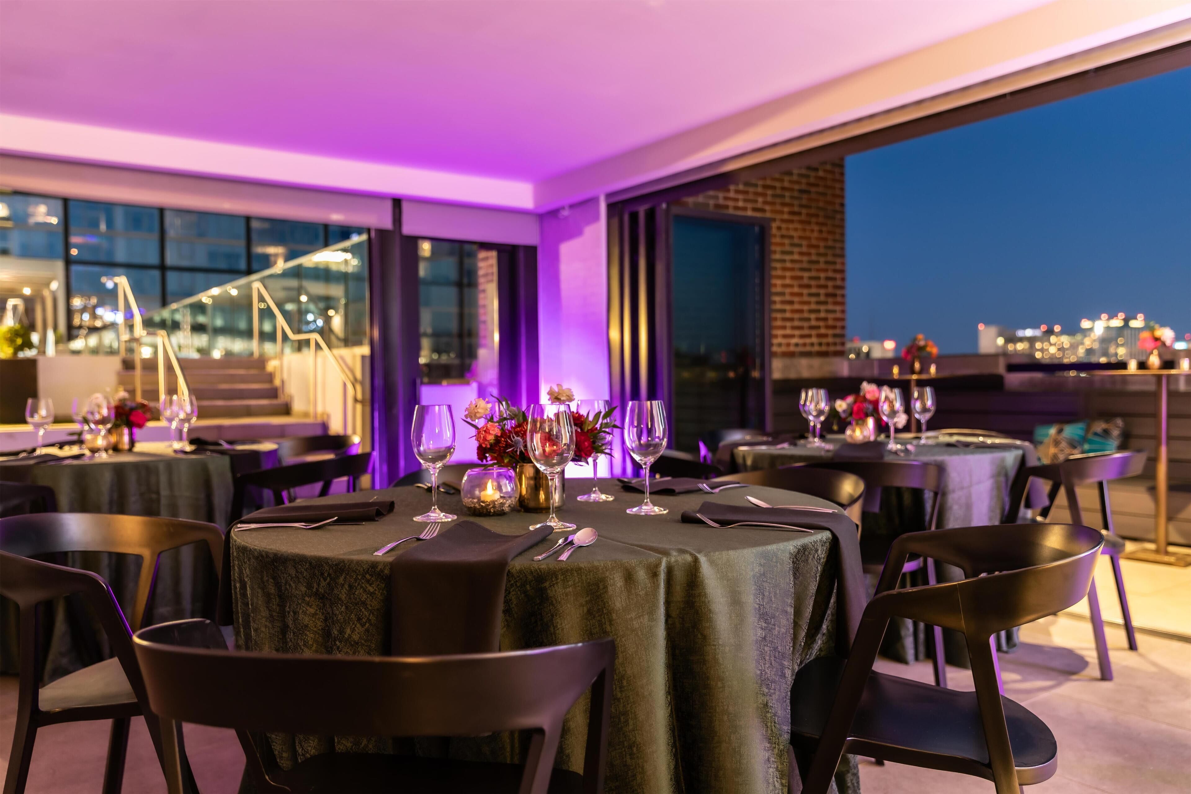 A rooftop dining area with tables set for two, featuring a city view at dusk.