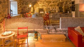 Una habitación rústica con paredes de piedra, una mesa de comedor de madera, sillas y sofá rojos, y una zona de cocina con refrigerador y horno.