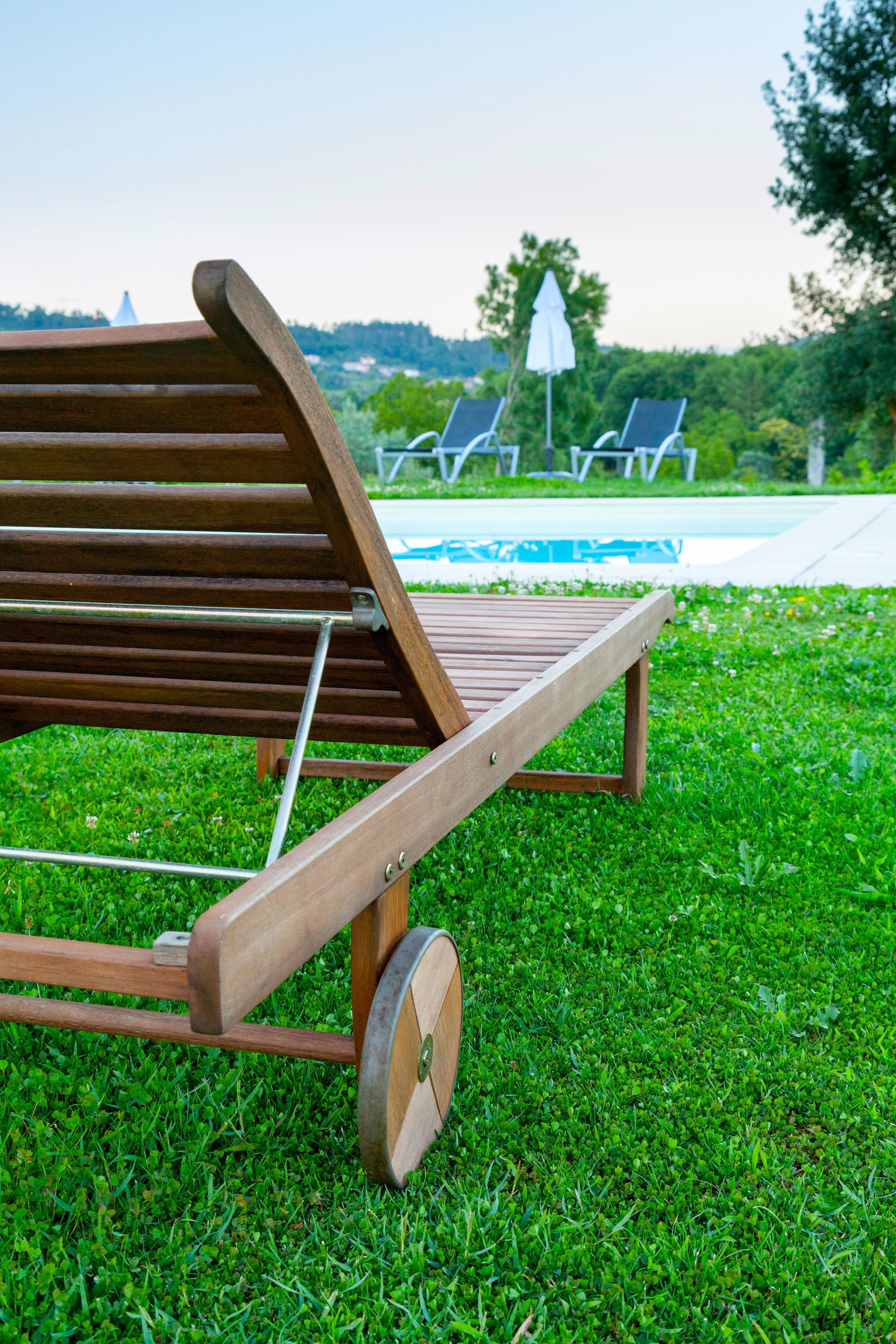 Ein Holz-Loungesessel mit Metallgestell, platziert auf einer Rasenfläche mit Pool und Sonnenschirmen im Hintergrund.