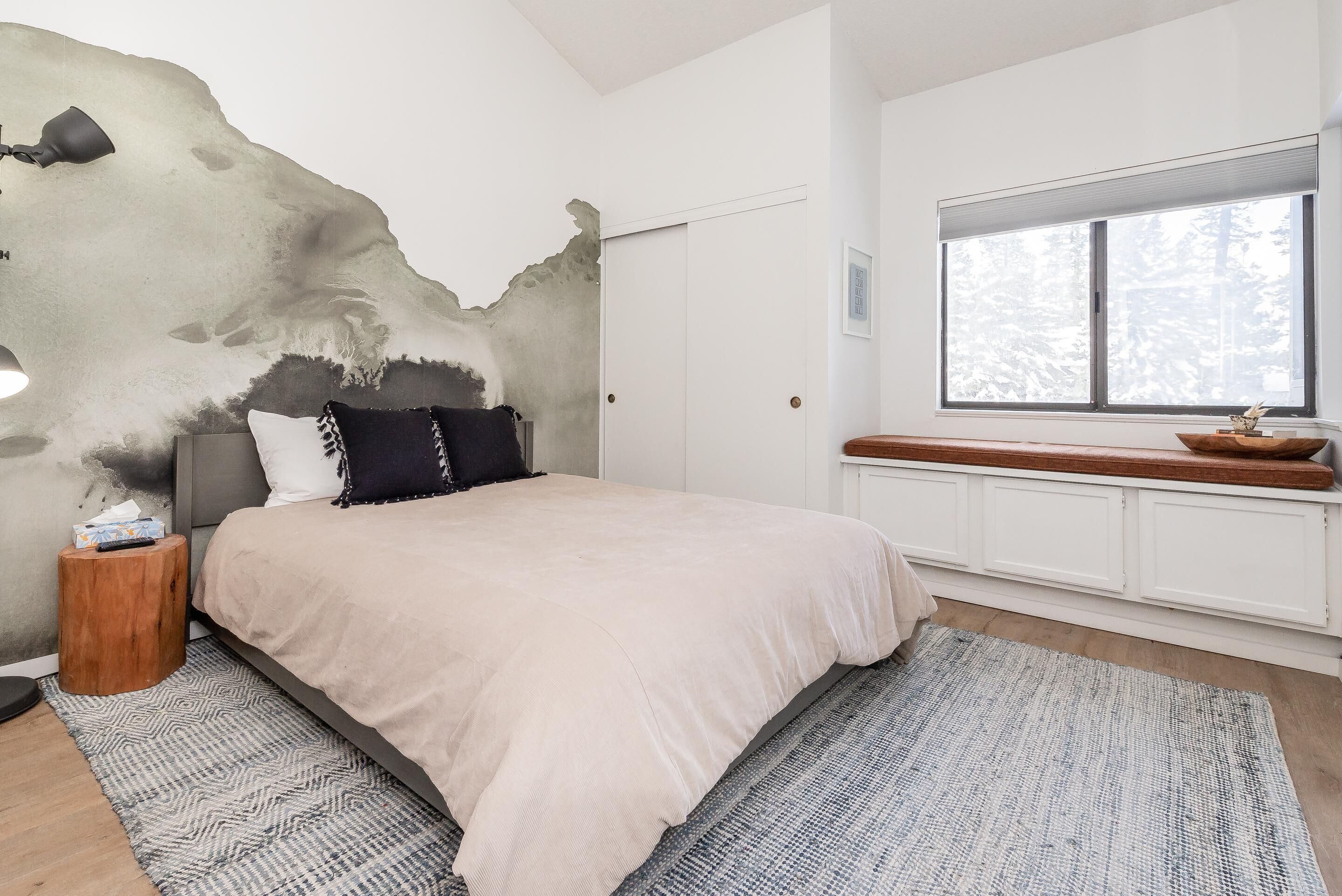 Un dormitorio con una cama grande, una mesita de noche, una pared con textura especial, un ventanal con vistas y un banco de madera junto a la ventana.