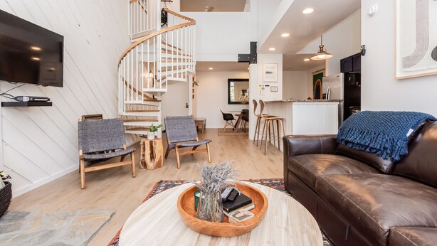 A spacious living area with a wooden staircase, a brown leather sofa, and a round wooden coffee table with books and a vase. - Crestview B44 (Mammoth Lakes)