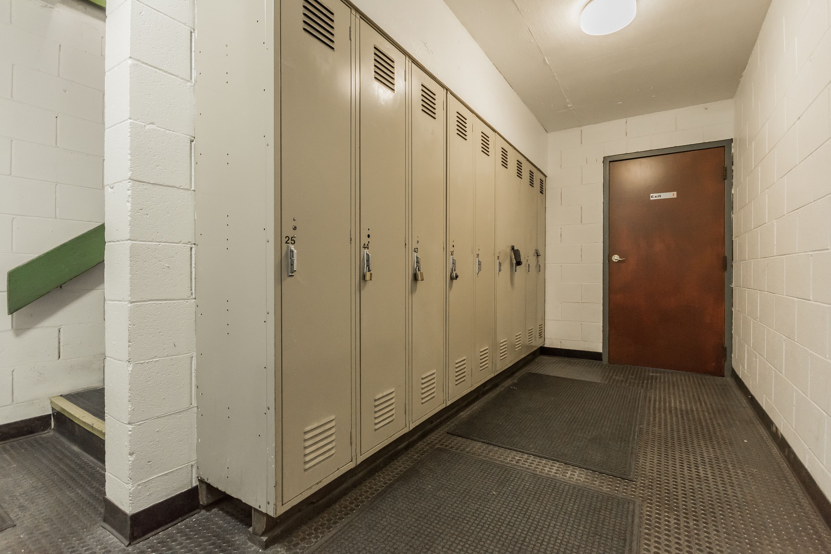A row of beige lockers with numbers on the doors, a green handrail, and a brown door at the end.