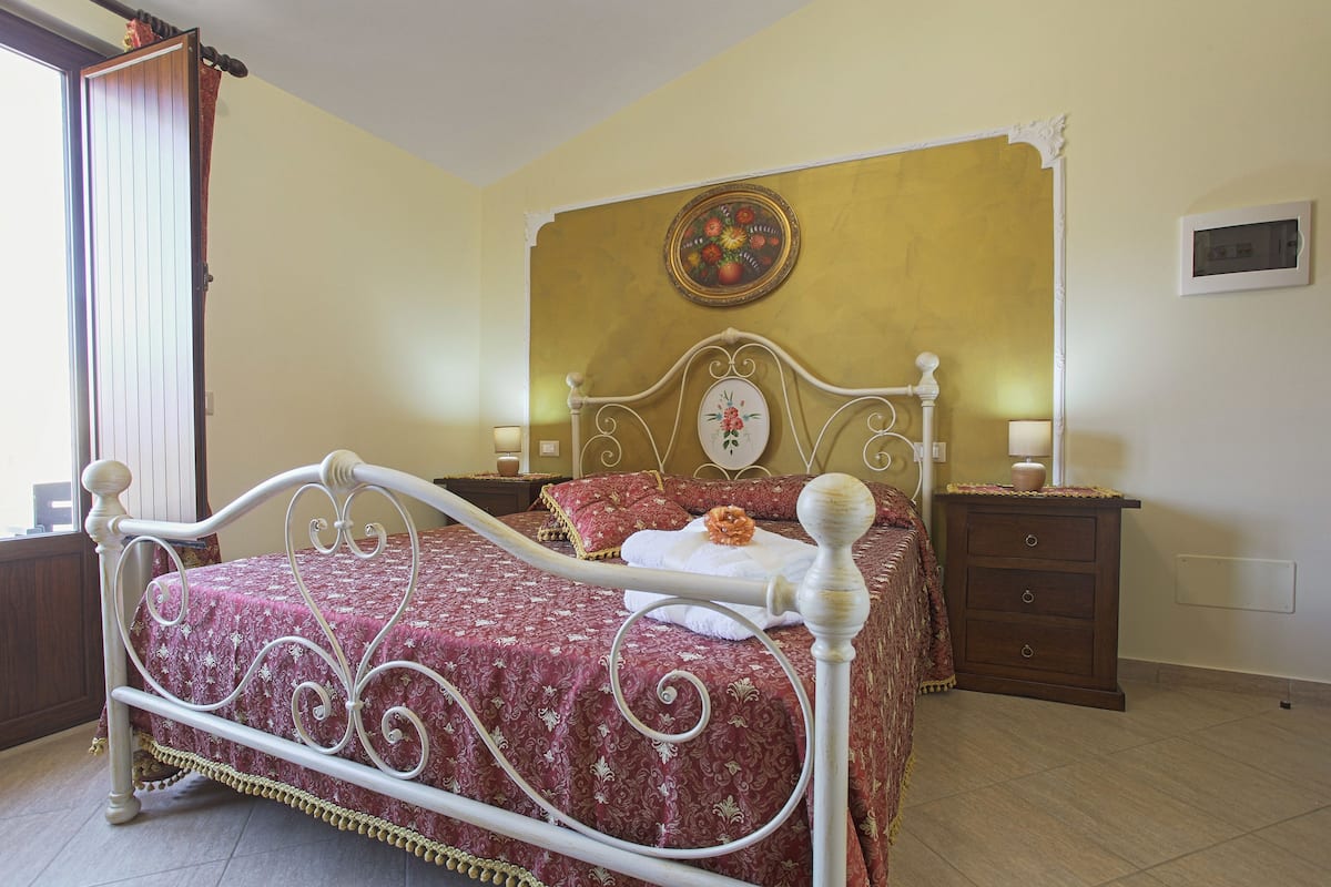 Un letto con struttura in metallo bianco, copripiumino rosso a fantasia e un cuscino decorativo. È presente un comodino con una lampada e una sveglia a muro.