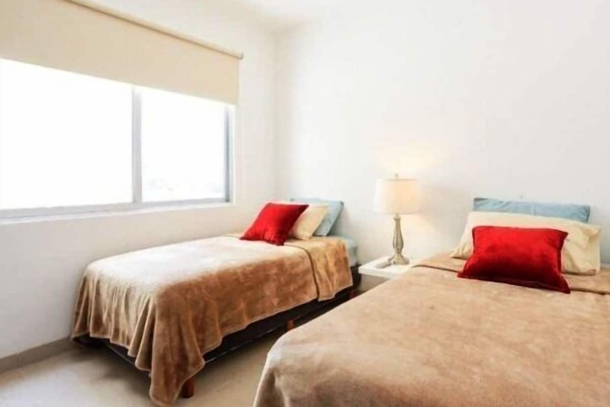 A room with two beds, each with a red pillow, a bedside lamp, and a window with a beige blind.