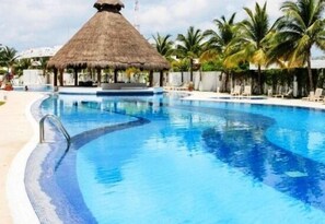 A swimming pool with a thatched-roof structure, palm trees, and lounge chairs. - Condo Esmir (Cancun)