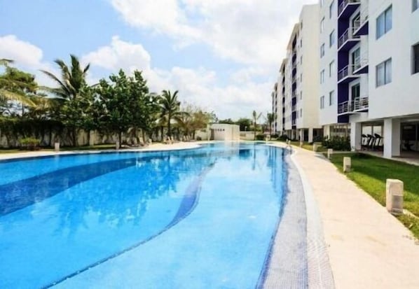 A swimming pool surrounded by a building with balconies, palm trees, and a clear sky.