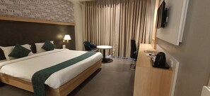 A hotel room with a large bed, a desk, a chair, a small table, a lamp, and a TV mounted on the wall. - Hotel Gopalas Residency (Bhiwandi)