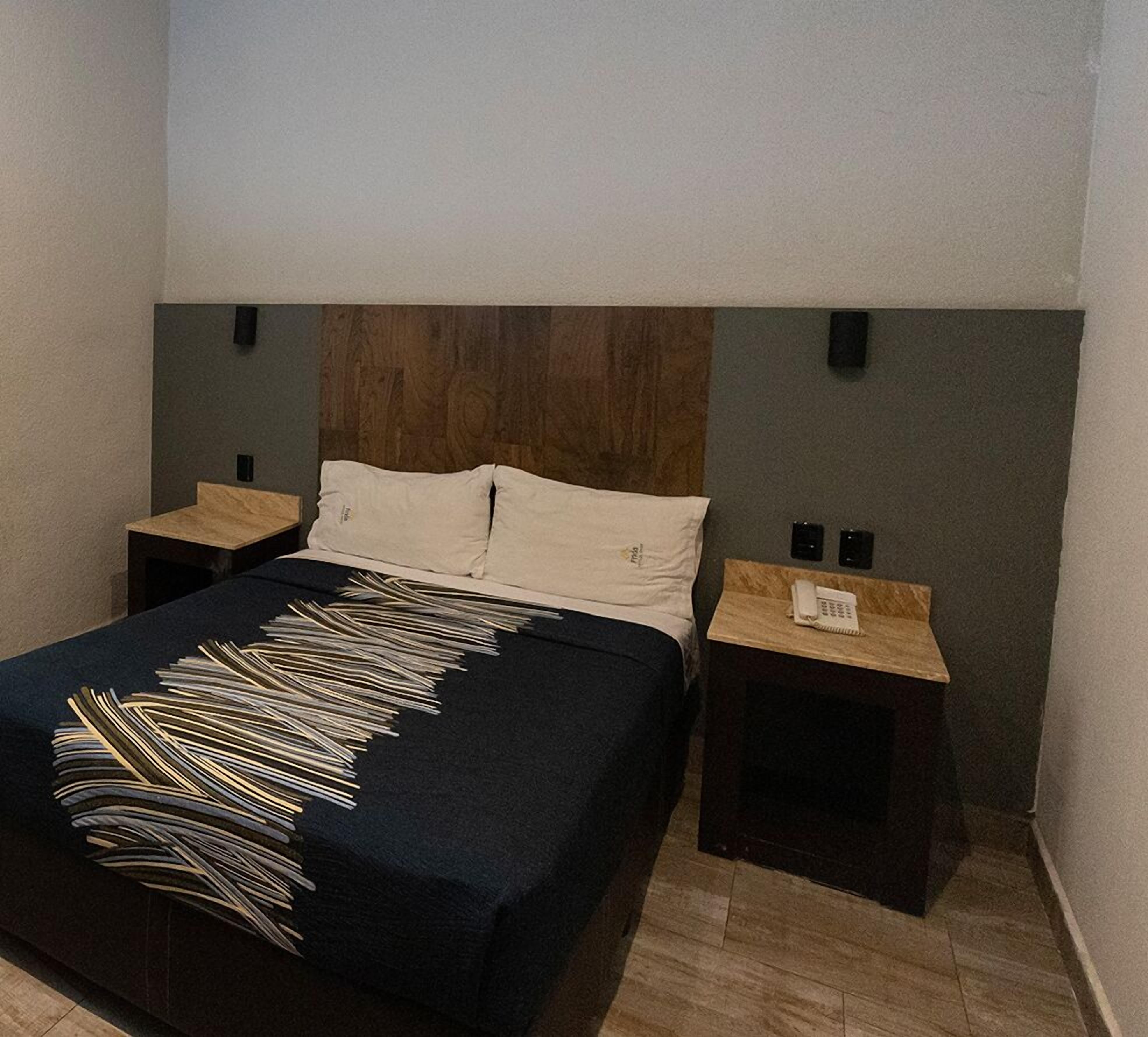 Habitación de hotel con cama, dos mesitas de noche, cabecero de madera y un teléfono sobre una de las mesitas.